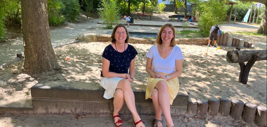 Kita Leiterin Susan Nestripke und Carolin Cezanne sitzend lächelnd auf einem Baumstamm im Außenbereich der Kita V