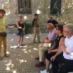 Bildungsfahrtteilnehmer und Stadtführer im Schatten sitzend und stehend vor einer alten Kirchenmauer.