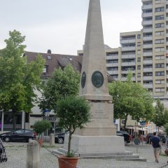 auf dem gepflasterten Platz sieht man einen Obelisken stehen, welcher von Bäumen eingerahmt ist.