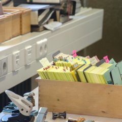 Ein brauner Holzkasten gefüllt mit gelben Karteikarten und mit bunten kleinen Fahnen versehen. 