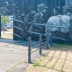 grau gemalte Mauer mit Menschen an der  ehemaligen Schlachthoframpe und im Vordergrund Eisengitter.