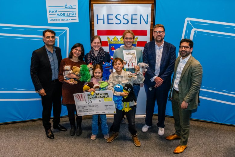Gruppenfoto vor dem hessischen Wappen. von links nach rechts hinten: 1. Kreisbeigeordneter Adil Oayn, drei Erwachsene von der Schule, Bürgermeister Karsten Groß, Verkehrsminister Kaweh Mansoori, vorne zwei Kinder mit großem symbolischen Scheck über 750€