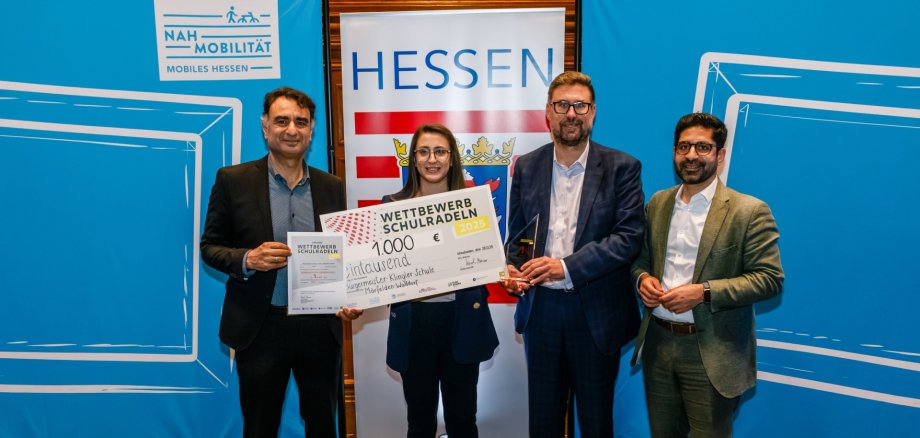 Gruppenfoto vor dem hessischen Wappen. von links nach rechts: 1. Kreisbeigeordneter Adil Oayn, eine Frau von der Schule hält großen symbolischen Scheck über 1000 €, Bürgermeister Karsten Groß, Verkehrsminister Kaweh Mansoori.