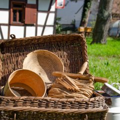 Weidenkorb vor altem Schuldhaus befüllt mit Holzsieb, Brotbackkorb und an der rechten Seite stehende weiße Milchkanne