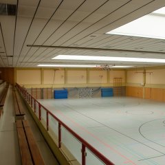 linke Seite lange Holzbänke für die Besucher und auf der rechten Seite Hallenboden mit roten, scharzen und blauen Linien sowie einem an der gegenüberliegenden Wand stehenden Tor