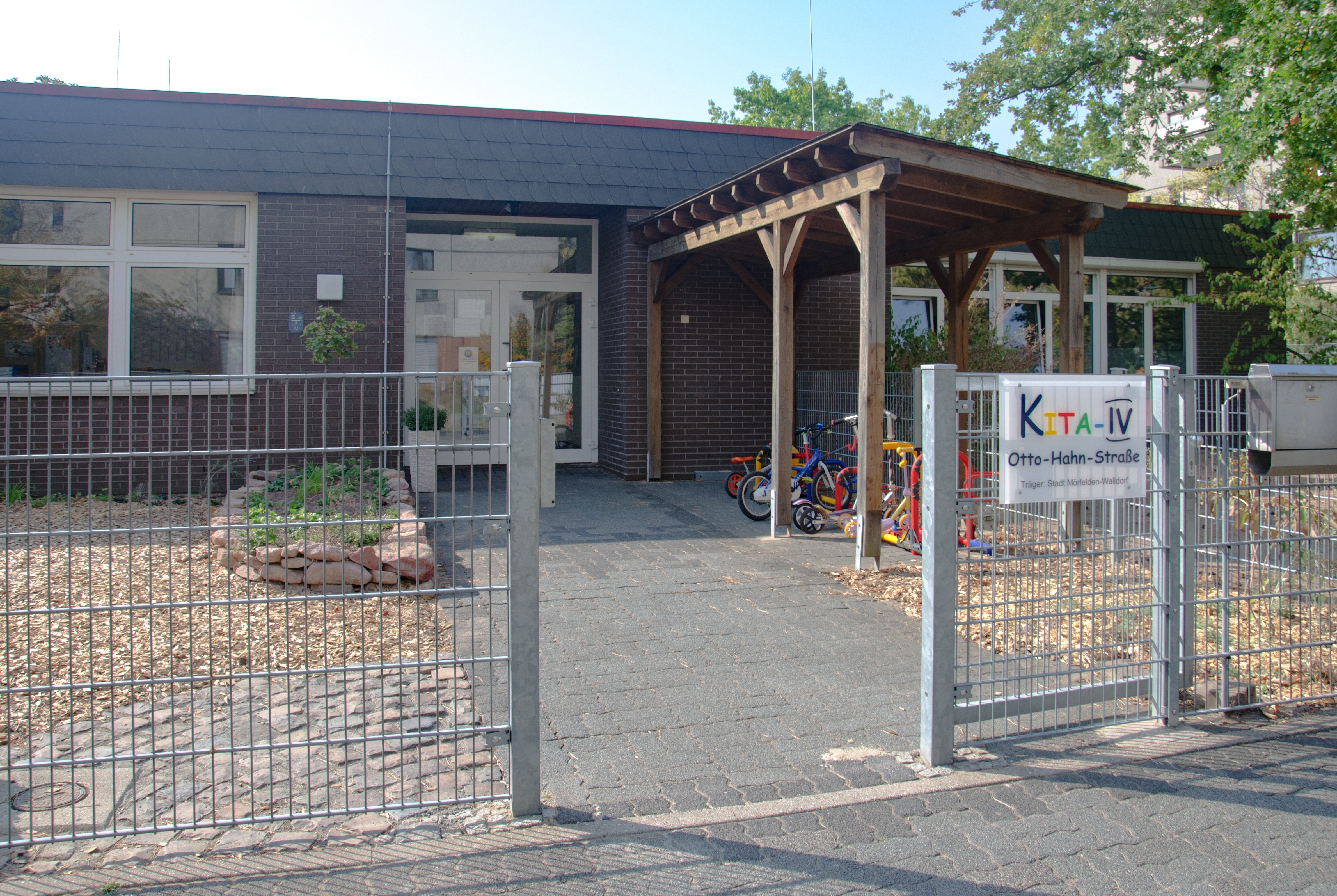 Eine weiße Eingangstür und auf der rechten Seite des Gebäudes. Davor eine Holzüberdachung für die Kinderfahrräder und ein buntes Schild mit der Aufschrift Kita 4.