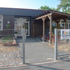 Eine weiße Eingangstür und auf der rechten Seite des Gebäudes. Davor eine Holzüberdachung für die Kinderfahrräder und ein buntes Schild mit der Aufschrift Kita 4.