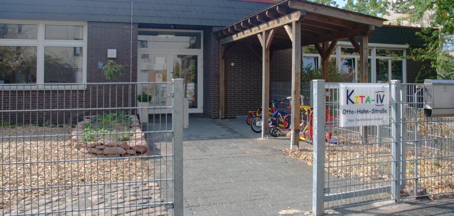Eine weiße Eingangstür und auf der rechten Seite des Gebäudes. Davor eine Holzüberdachung für die Kinderfahrräder und ein buntes Schild mit der Aufschrift Kita 4.