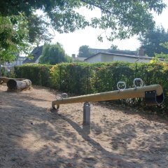 Eine  Holzwippe mit vier Griffen vor einer grünen Hecke.