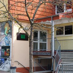 Eine große breite Treppe führt zum Eingang. An der Hauswand sieht man ein großes gesprüthes Wandbild.
