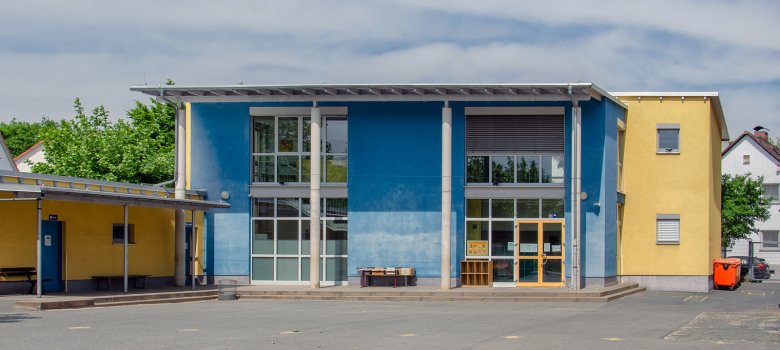 Blaues Schulgebäude mit großen weißen Fensterfronten und davor ein großer geteerter Schulhof.