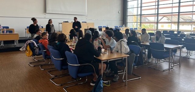 Gruppe von ca. 20 Jugendlichen sitzen an zwei Tischen im Stadtverordnetensitzungssaal. Im Hintergrund stehen zwei Frauen und Bürgermeister Groß und sprechen zu den Austauschschülern.