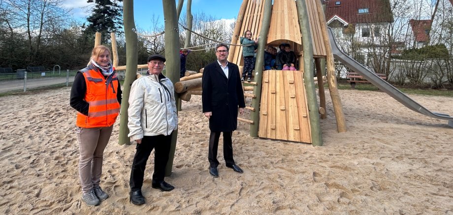 Neues Klettergerüst aus Holz in Form eines Indianerzelts mit Rutsche. Im Vordergrund im Sand stehen Susanne Görlich (Bauhof), Georg Germann (Magistrat) und Karsten Groß (Bürgermeister).