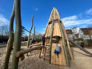 Neues Klettergerüst aus Holz in Form eines Indianerzelts mit Rutsche. Ein Kind klettert an der Außenwand hoch.