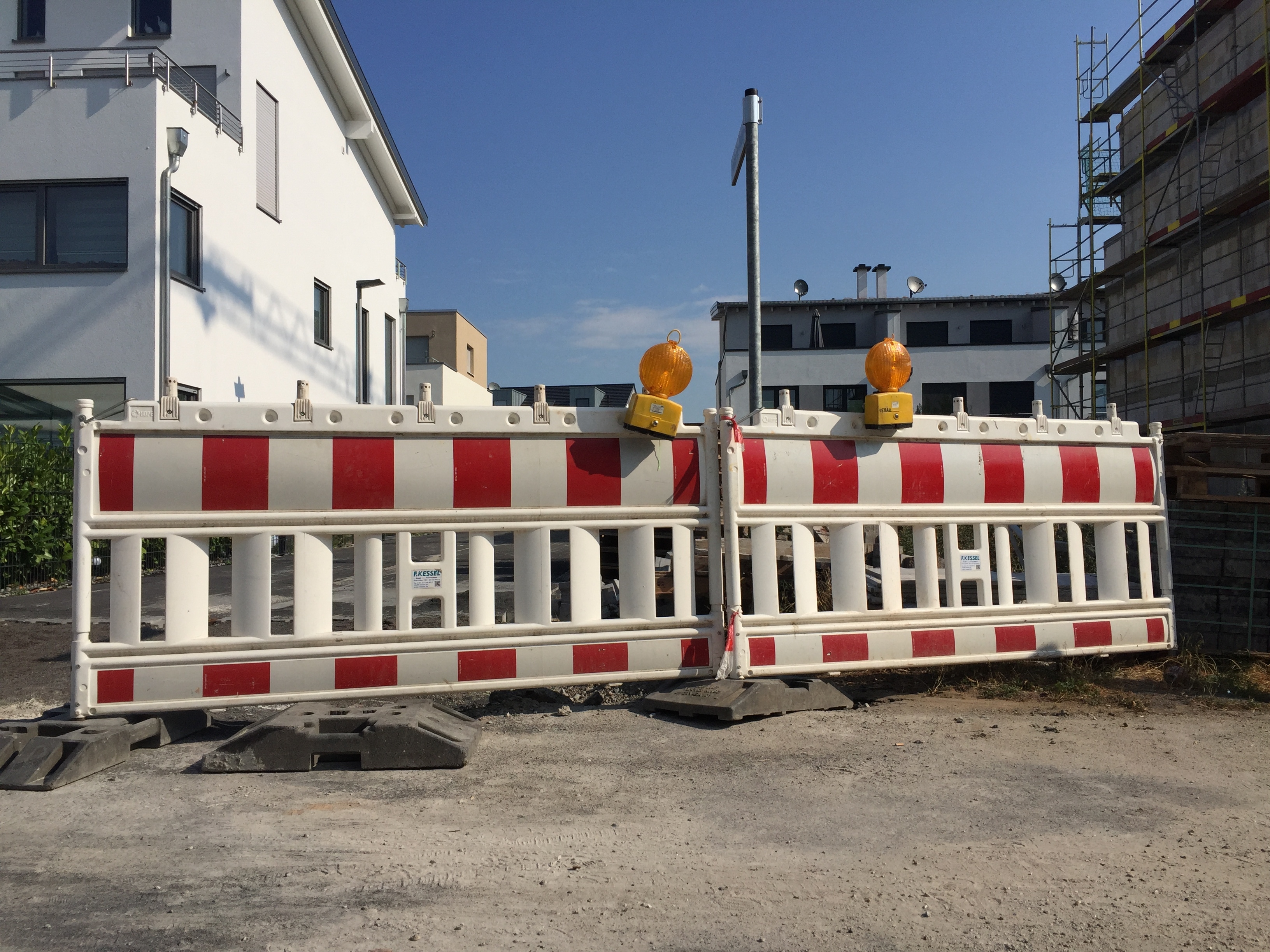 Zwei rot-weißgestreifte Straßenabsperrungen stehen nebeneinander, darauf befinden sich jeweils eine gelbe Warnleuchte. Hinter der Absperrung sieht man Wohnblocks.