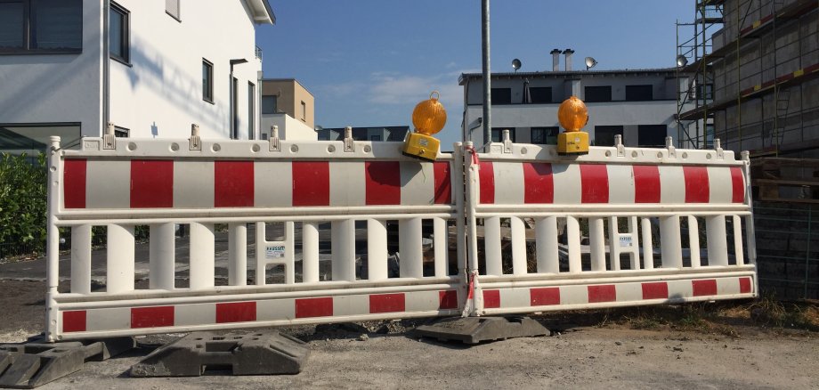 Zwei rot-weißgestreifte Straßenabsperrungen stehen nebeneinander, darauf befinden sich jeweils eine gelbe Warnleuchte. Hinter der Absperrung sieht man Wohnblocks.
