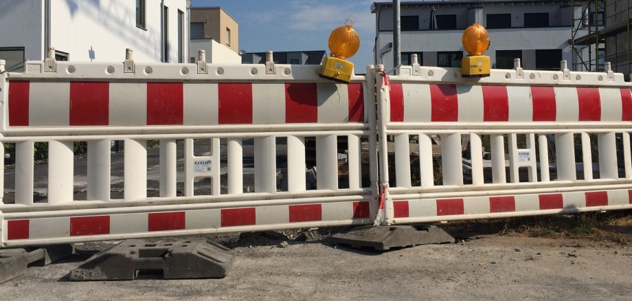 Zwei rot-weißgestreifte Straßenabsperrungen stehen nebeneinander, darauf befinden sich jeweils eine gelbe Warnleuchte. Hinter der Absperrung sieht man Wohnblocks.  
