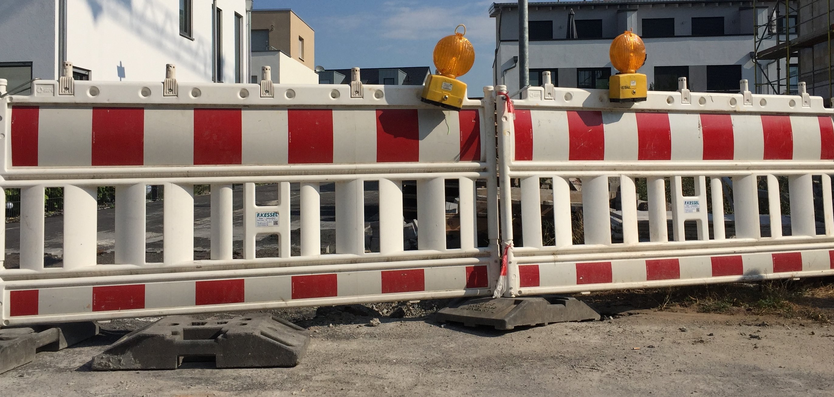 Zwei rot-weißgestreifte Straßenabsperrungen stehen nebeneinander, darauf befinden sich jeweils eine gelbe Warnleuchte. Hinter der Absperrung sieht man Wohnblocks.