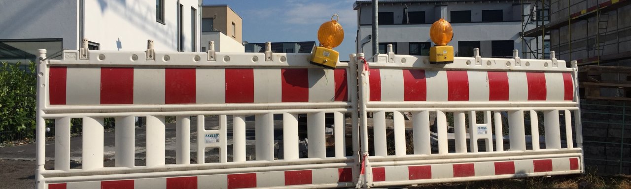 Zwei rot-weißgestreifte Straßenabsperrungen stehen nebeneinander, darauf befinden sich jeweils eine gelbe Warnleuchte. Hinter der Absperrung sieht man Wohnblocks.