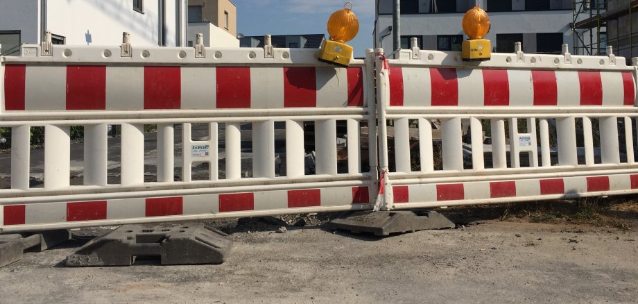 Zwei rot-weißgestreifte Straßenabsperrungen stehen nebeneinander, darauf befinden sich jeweils eine gelbe Warnleuchte. Hinter der Absperrung sieht man Wohnblocks.
