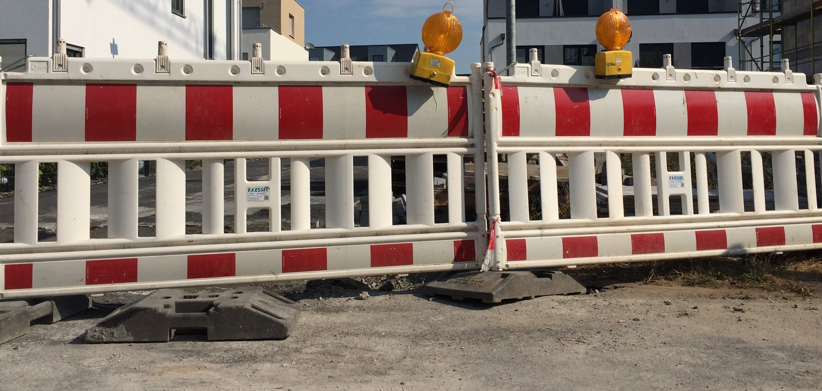 Zwei rot-weißgestreifte Straßenabsperrungen stehen nebeneinander, darauf befinden sich jeweils eine gelbe Warnleuchte. Hinter der Absperrung sieht man Wohnblocks.