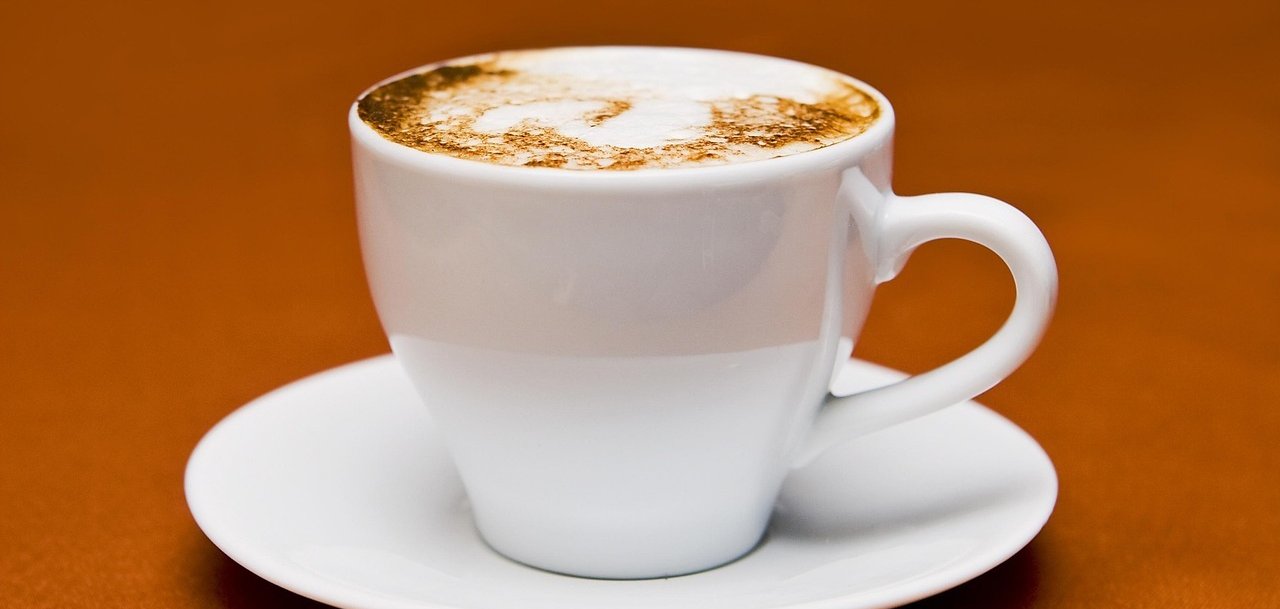Weiße Tasse mit Untertasse und Cappuccino darin auf braunen Untergrund.