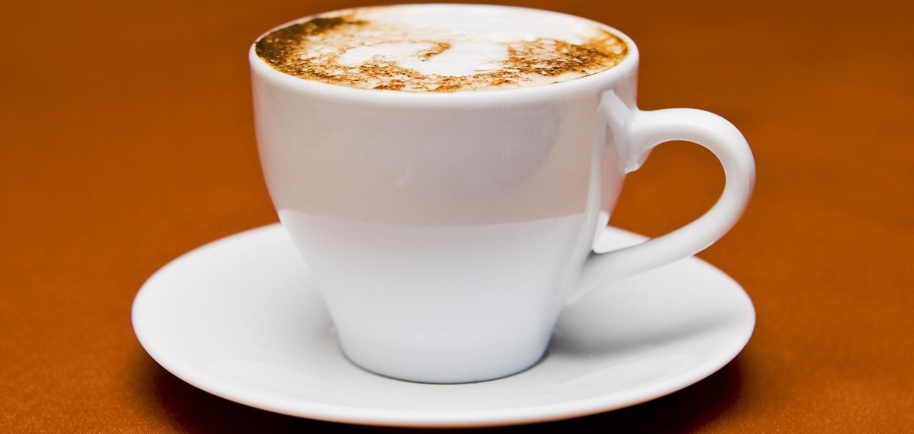 Weiße Tasse mit Untertasse und Cappuccino darin auf braunen Untergrund.