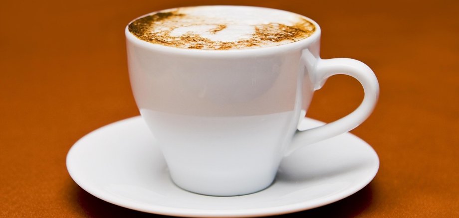 Weiße Tasse mit Untertasse und Cappuccino darin auf braunen Untergrund.