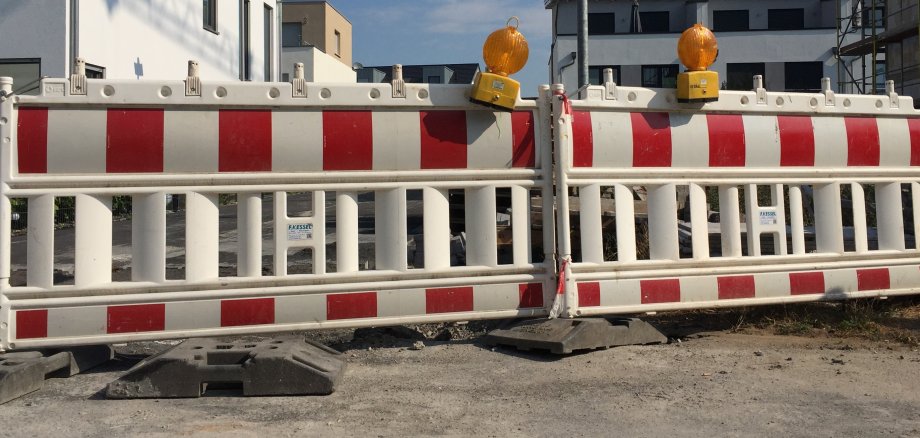 Zwei rot-weißgestreifte Straßenabsperrungen stehen nebeneinander, darauf befinden sich jeweils eine gelbe Warnleuchte. Hinter der Absperrung sieht man Wohnblocks.