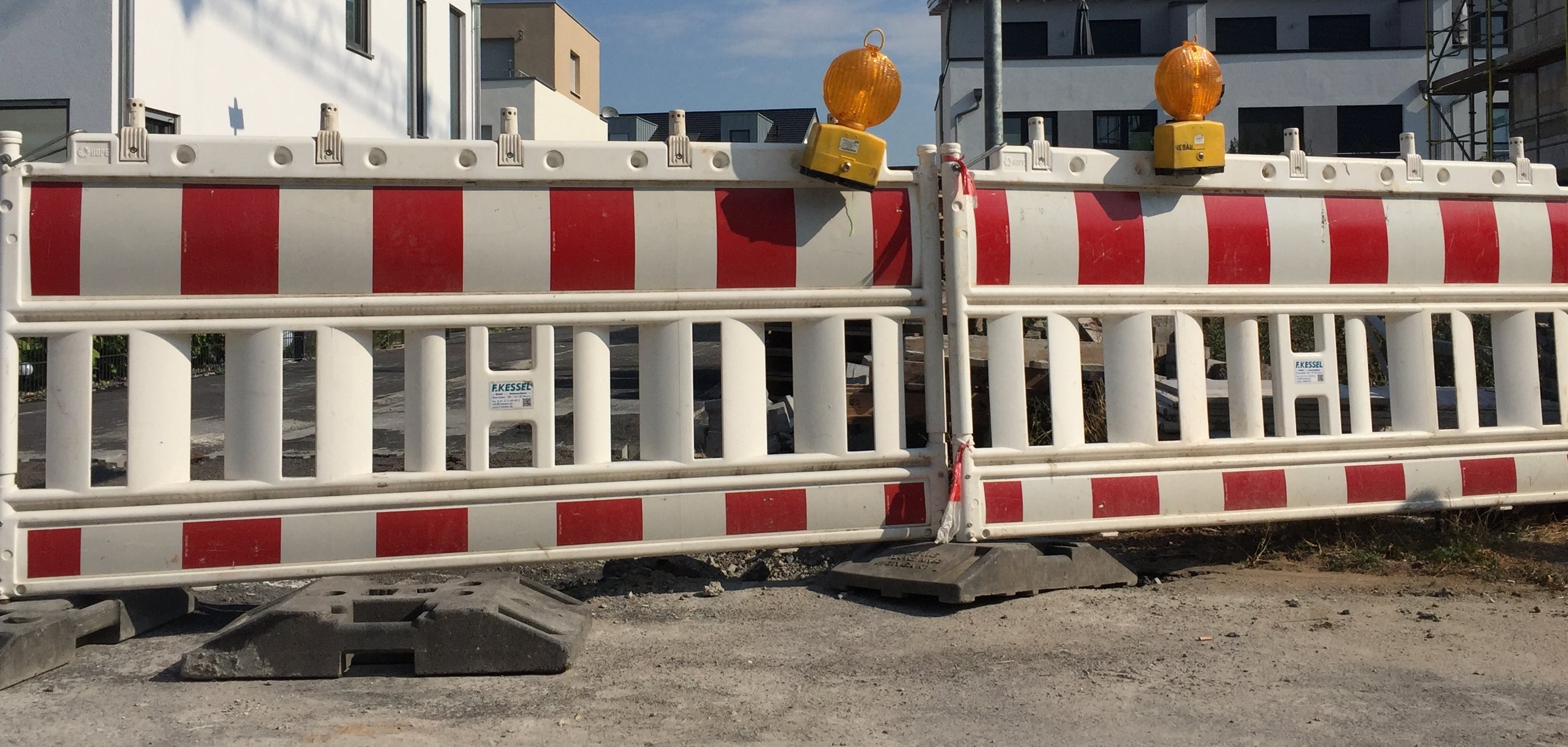 Zwei rot-weißgestreifte Straßenabsperrungen stehen nebeneinander, darauf befinden sich jeweils eine gelbe Warnleuchte. Hinter der Absperrung sieht man Wohnblocks.  