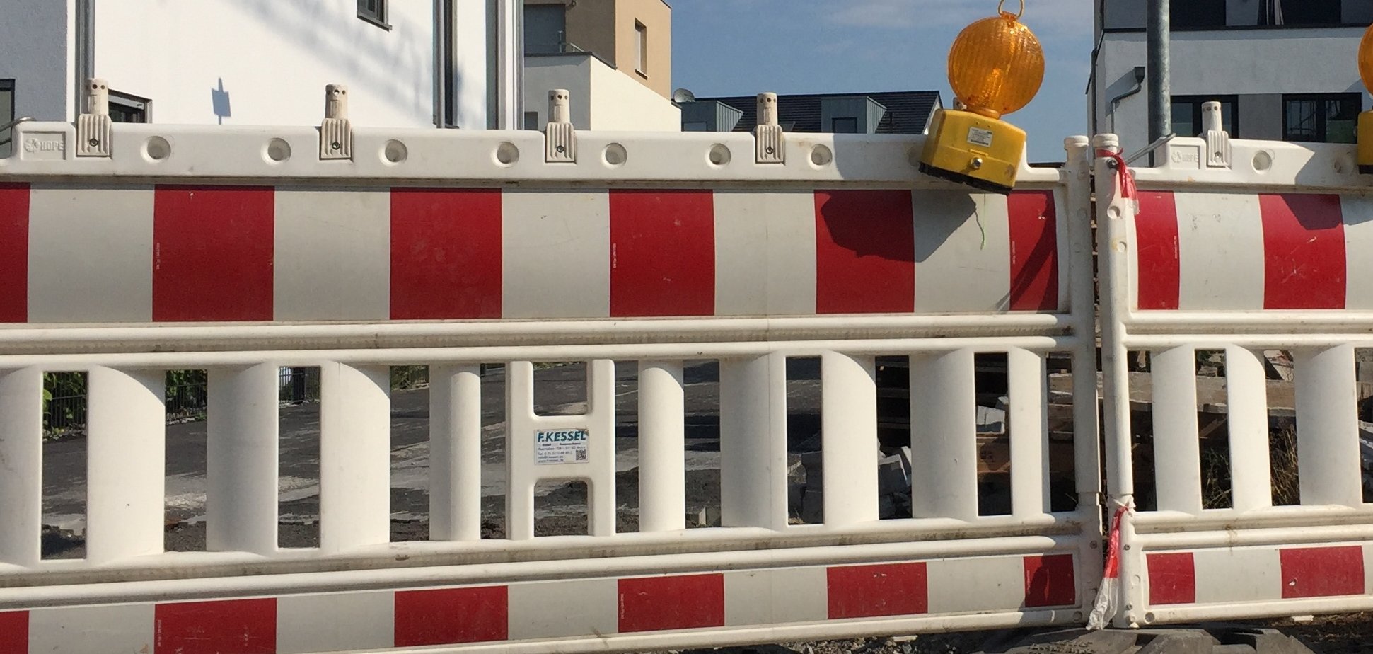 Zwei rot-weißgestreifte Straßenabsperrungen stehen nebeneinander, darauf befinden sich jeweils eine gelbe Warnleuchte. Hinter der Absperrung sieht man Wohnblocks.