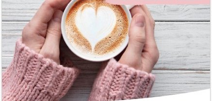 Tasse mit Cappuccino und aufgeschäumter Milch mit Herzmuster gehalten in zwei Händen.