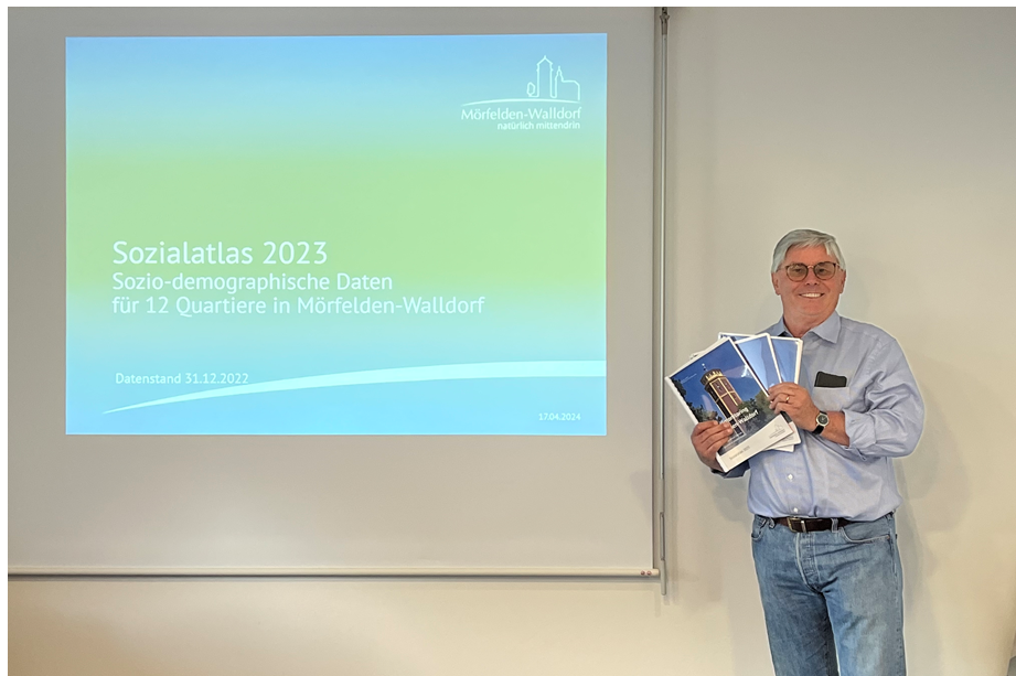 Heimo Boschert steht mit dem Sozialatlas lächelnd rechts vor einer PowerPointPräsentation, die auf eine Leinwand projehziert wurde.