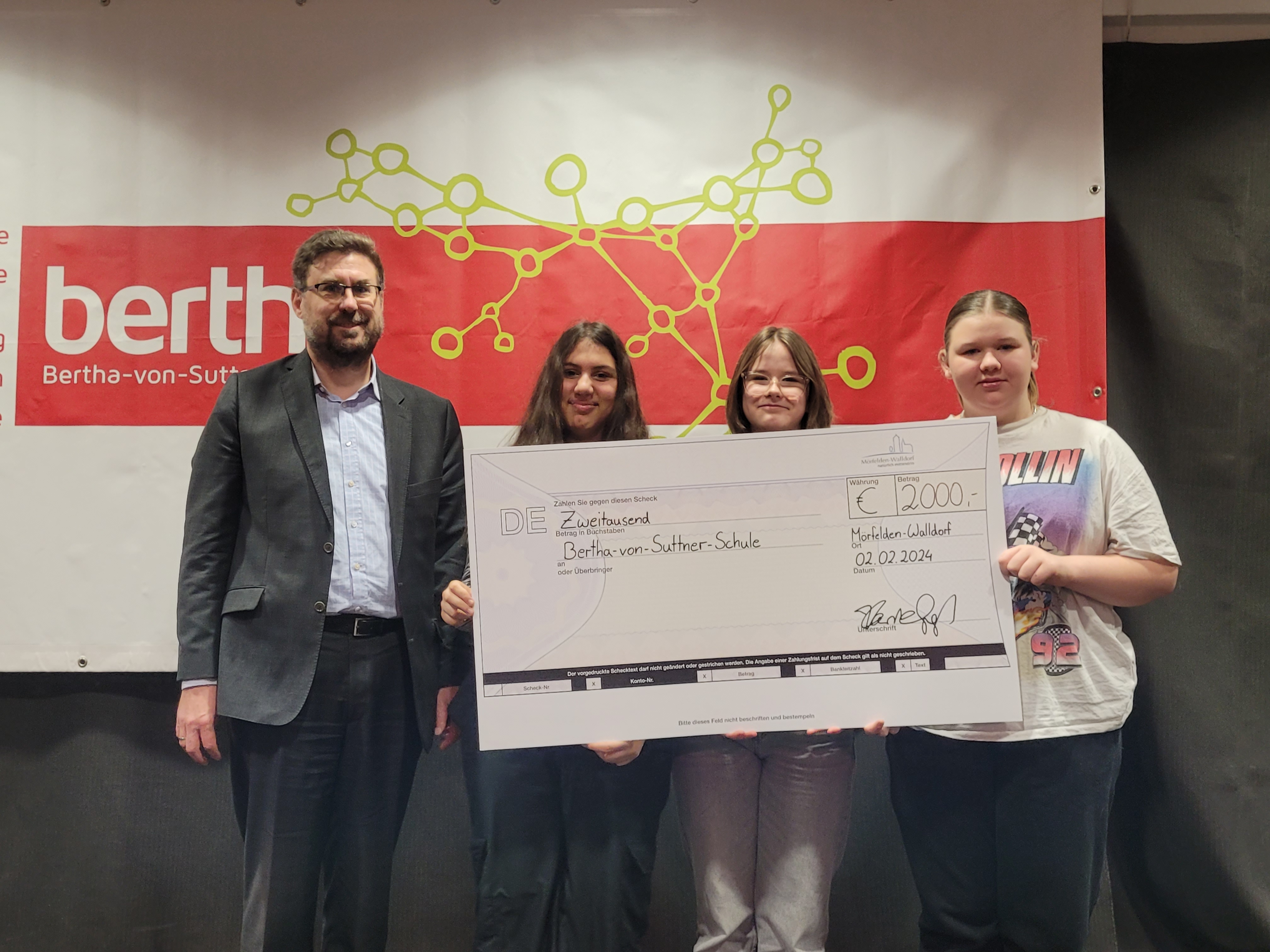 Karsten Groß und drei 7 Klässerinnen halten einen großen Scheck mit der Aufschrift 2.000 Euro. Hinter ihnen ist ein Banner der bertha zu sehen.
