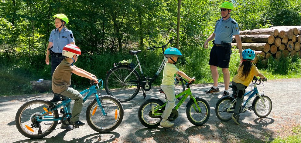Kinder fahren mit Fahrrädern und Schutzhelmen an einer Polizistin und einem Polizisten vorbei