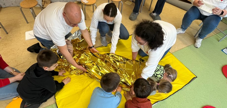 drei Kinder und drei Mitarbeiter (ein Mann, zwei Frauen) wickeln zwei Kinder in eine DRK-Decke