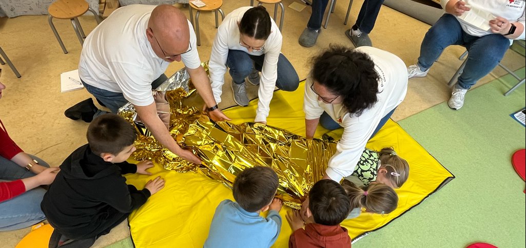 drei Kinder und drei Mitarbeiter (ein Mann, zwei Frauen) wickeln zwei Kinder in eine DRK-Decke