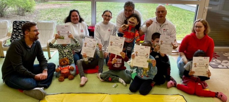 Kinder halten ihre Urkunden vor das Gesicht. Erster Stadtrat Karsten Groß sitzt daneben. Auf dem Bild zu sehen sind auch Mitarbeitende des Deutschen Roten Kreuzes