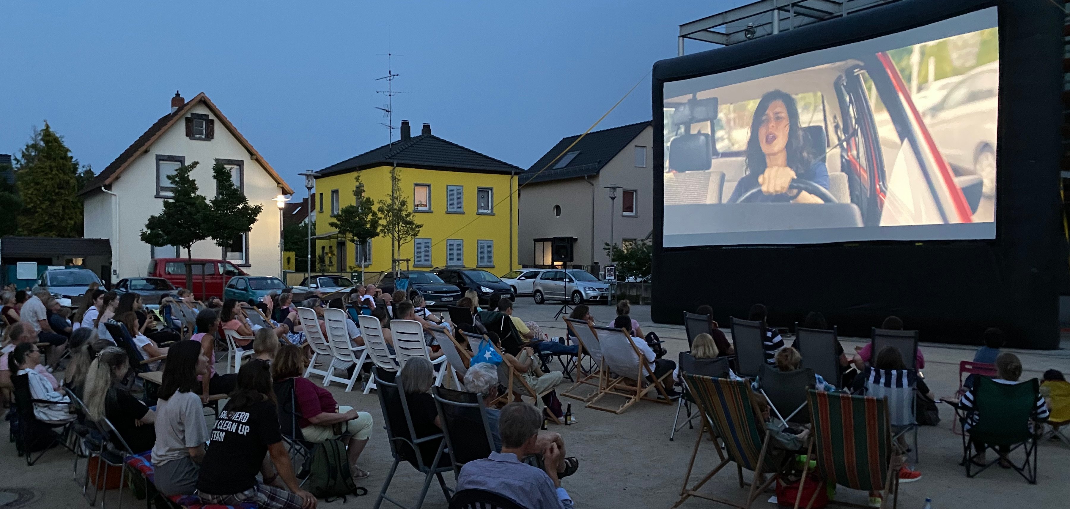 Zuschauer und Zuschauerinnen seitlich von hinten fotografiert schauen auf eine Kinoleinwand. im Hintergrund sind Häuser zu sehen.