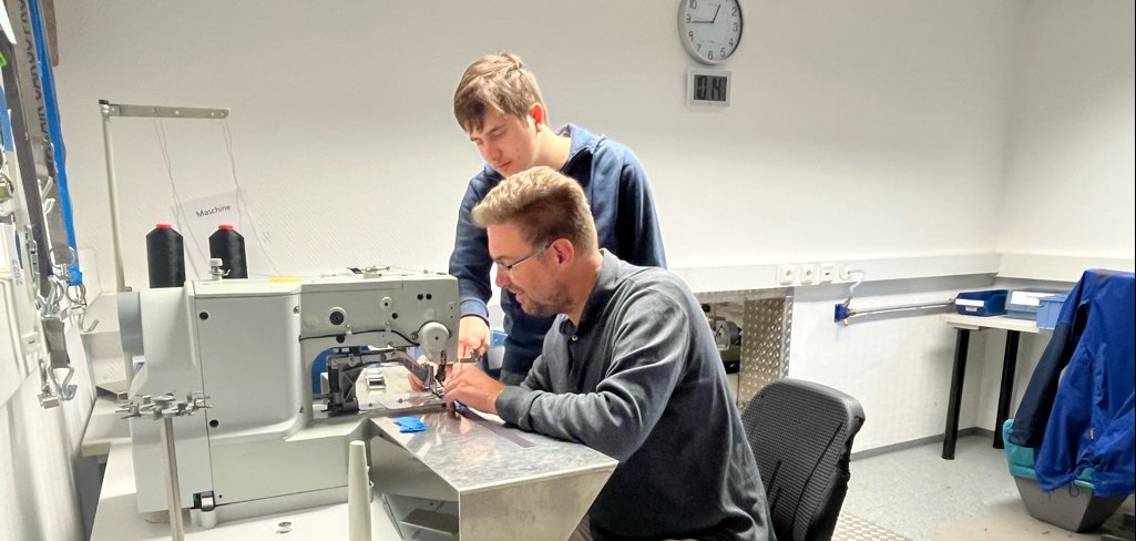 Karsten Groß an der Nähmschine. Links neben ihm steht Mitarbeiter Alexander. Sie schauen sich den genähten Gurt an