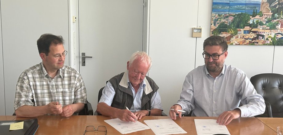 Archivar Torsten Petri, Rudi Hechler und Büregermeister Karsten Groß sitzen nebeneinander. Rudi Hechler unterschreibt den Schenkungsvertrag, der auf dem Tisch liegt. Karsten Groß lächelt, die anderen schauen auf den Vertrag