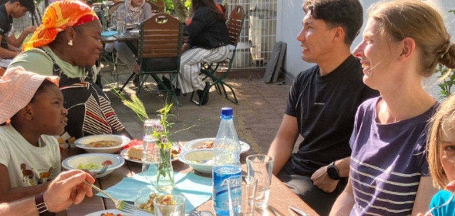 Eine Gruupe von Menschen verschiedener Haufarben sitzen lachend vor einem Tisch mit Essen