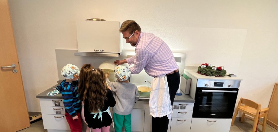 Erster Stadtrat Karsten Groß umringt von Kindern schüttet Gemüse in den Kochtopf, der auf dem Herd der Kinderküche steht