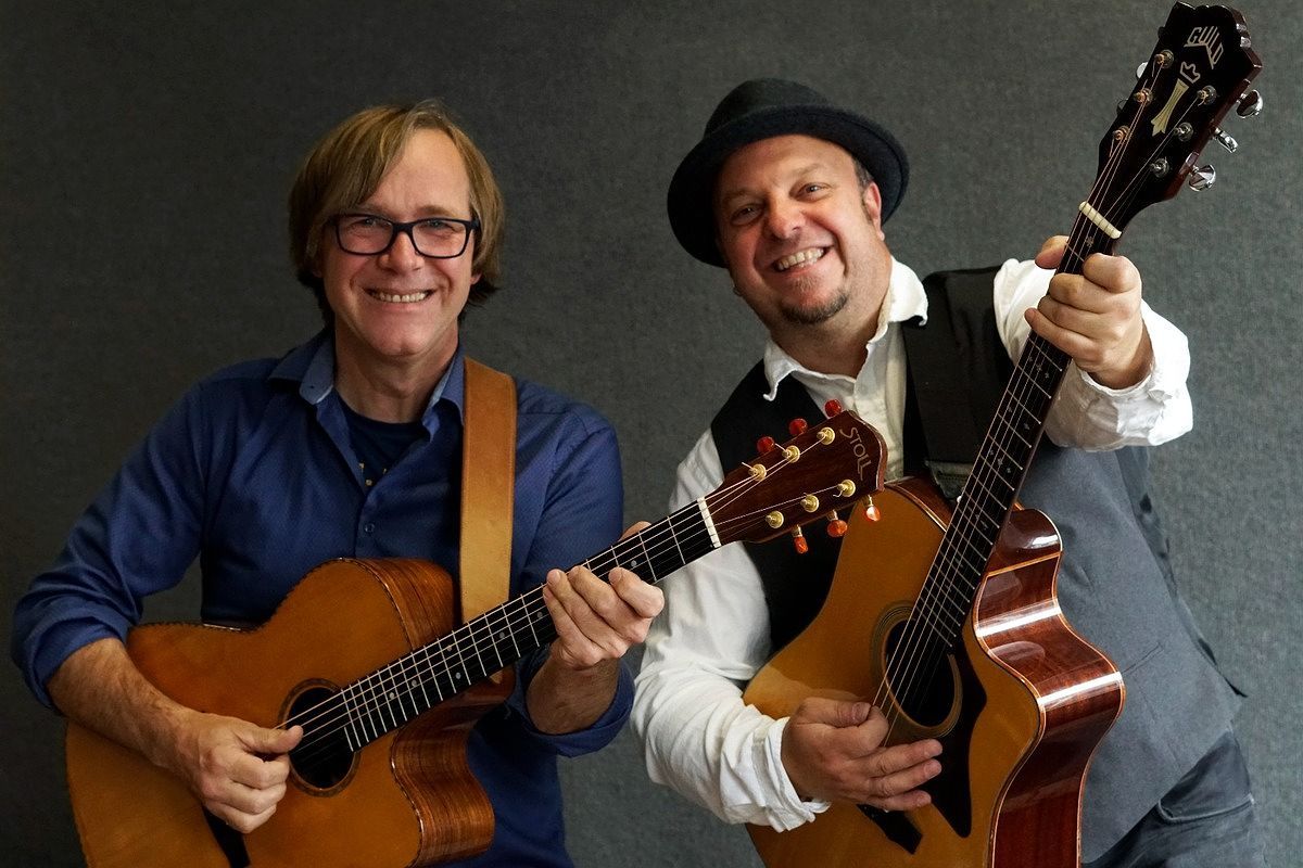 Beide Musiker halten Ihre Gitarren in der Hand und lächeln in die Kamera. Während der linke Mann mit der schwarzen Brille gerade steht, lehnt sich der rechte Mann mit Hut leicht nach hinten gelehnt in Richtung seines Bandkollegen.