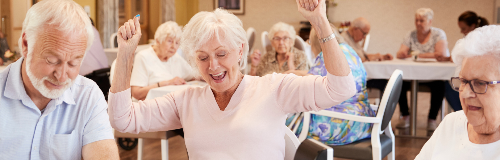 Design ohne Titel - Bingo Zu sehen ist ein Raum mit mehreren Seniorinnen und Senioren, die an Tischen sitzen. Eine Frau im Vordergrund hat beide Arme triumphierend in die Höhe gestreckt und lacht.