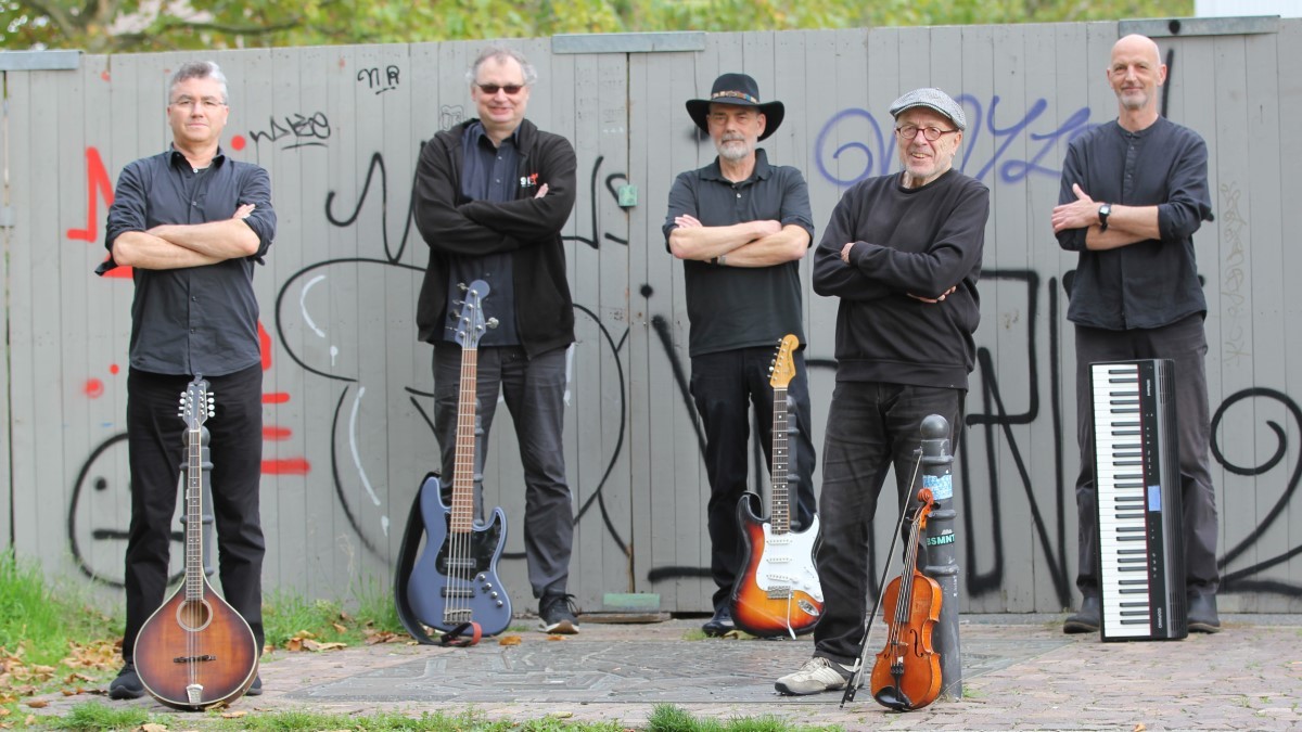 5 schwarzgekleidete Musiker stehen mit ihren Instrumenten locker aufgereiht vor einer grauen Mauer.