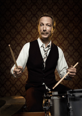 Der Künstler hält zwei Drumsticks in Händen und grinst in die Kamera