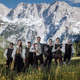 Sieben Musizierende in traditioneller Kleidung lächeln in die Kamera. Im Hintergrund sieht man eine Berglandschaft.