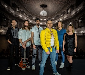 Die sechs Musiker:innen stehen vor dem Hintergrund eines großen Konzertsaales.